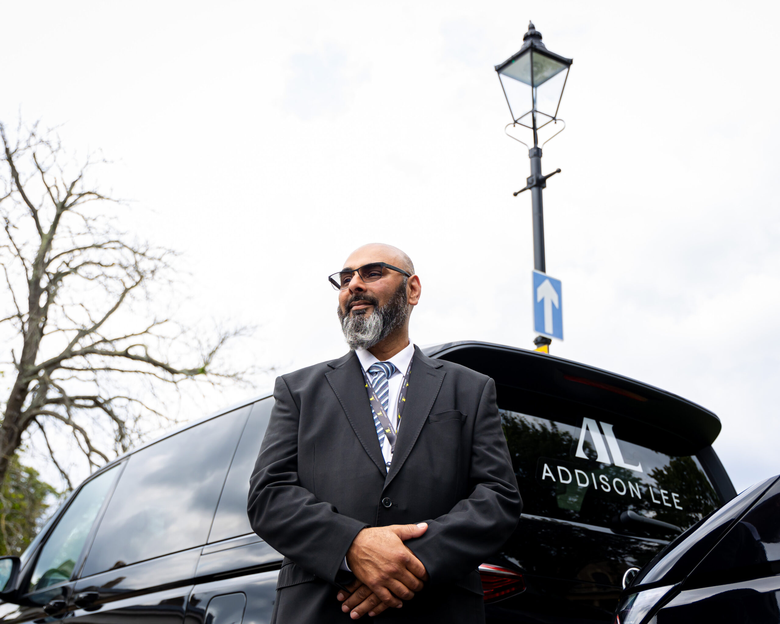 Driver wearing sunglasses stood behind an Addison Lee taxi with his arms crossed