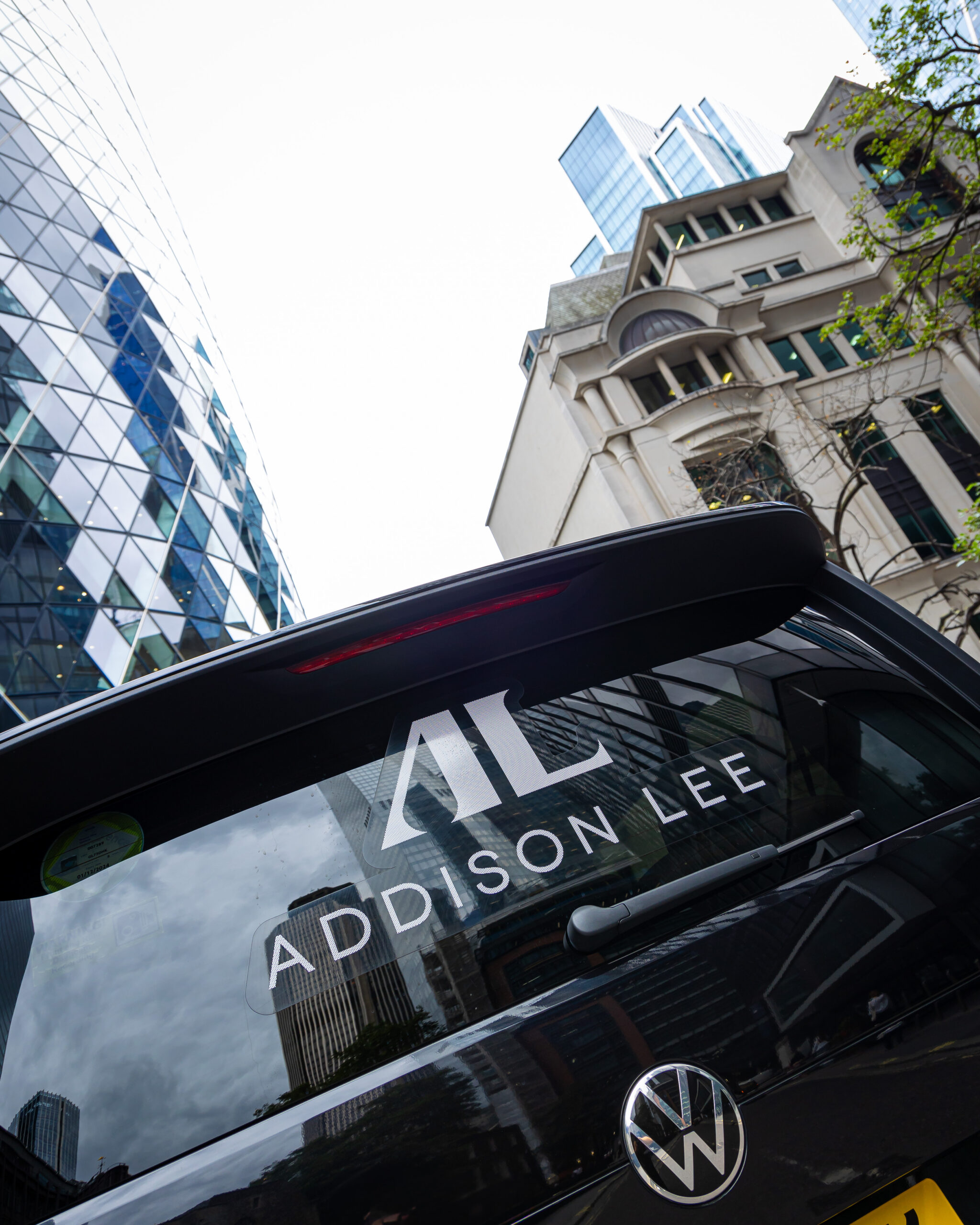 Close up of the back of an Addison Lee taxi with the gherkin and various London city skyscrapers in the background